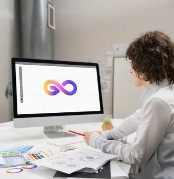 An Employee is sitting on a chair holding a pen in one hand and a mouse in another and designing a logo on a computer
